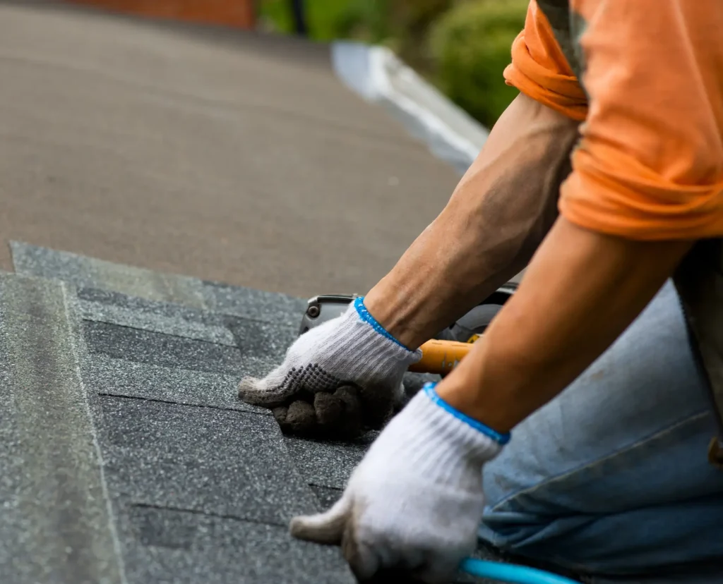 A construction worker wearing white gloves and an orange shirt installs shingles on a roof. The worker, from a Roofing Company in Arcadia LA, is using tools to secure the shingles in place. The close-up view shows the worker's hands and arms while focusing on the roofing task.