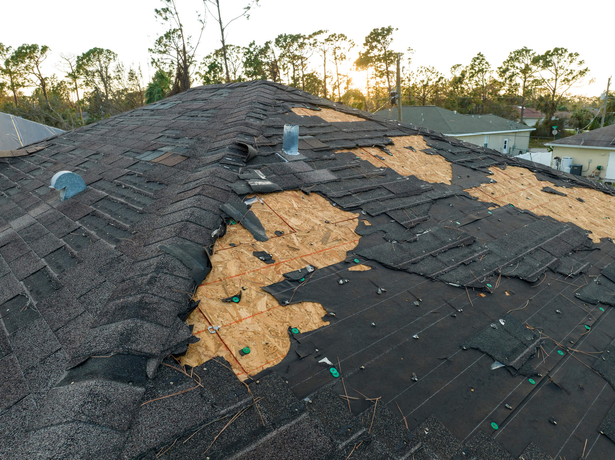 A roof with significant damage, showing torn and missing shingles, exposing the underlying wood and protective material. The area around the damage has scattered debris, and the background reveals some trees and houses at sunset—a stark reminder that maintenance can affect how long roofs last.