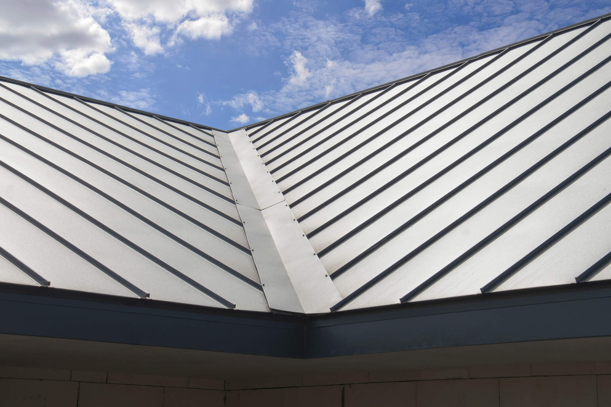 A close-up view of a modern metal roof with a ridge cap, against a backdrop of a partly cloudy blue sky. The roof features sleek, parallel lines running to the ridge, creating a clean and symmetrical appearance. What is your opinion of metal shingle roofing in such elegant designs?