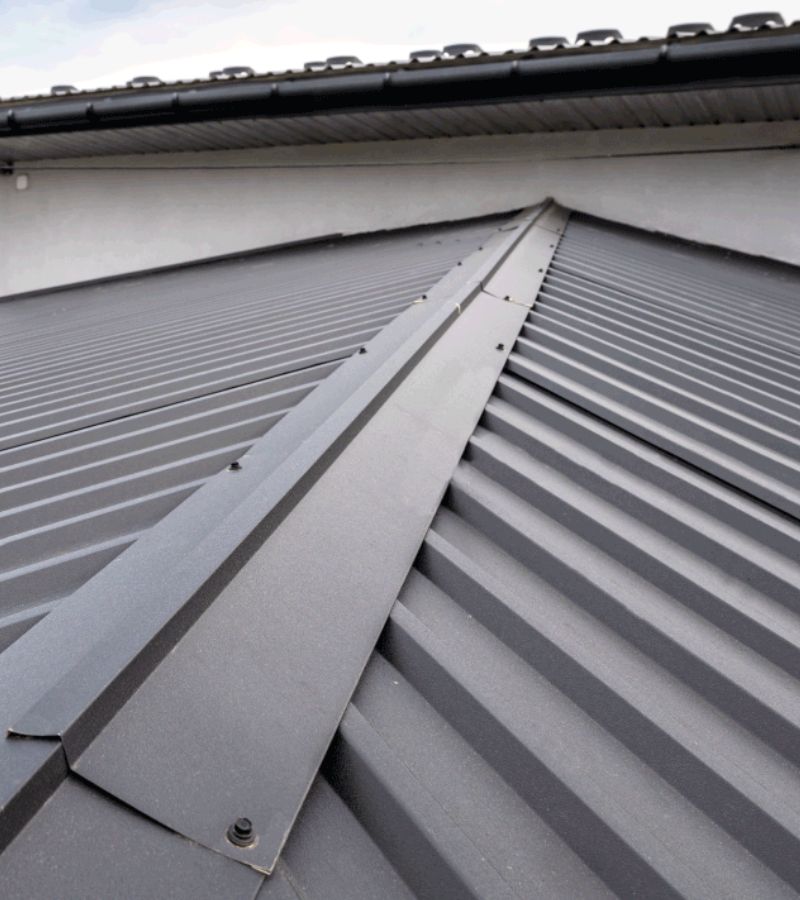 A close-up view of a building's roof, featuring a grey metal roof with a central ridge. The roof has corrugated panels and appears clean and well-maintained, highlighting the benefits of a metal roof over shingles. The edge of the roof includes a gutter system. Sky and wall of the building are visible in the background.