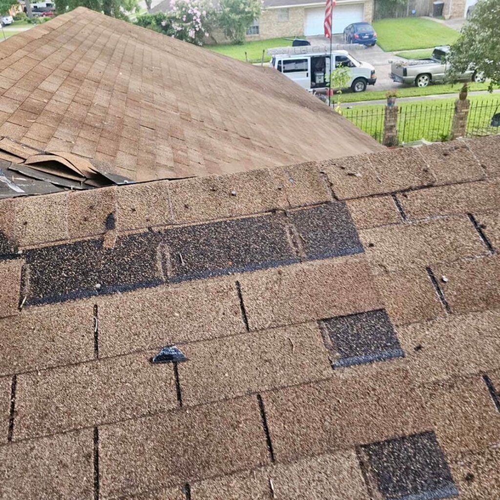 A rooftop with significant shingle damage displays missing and loose shingles, showing clear signs of roof damage. In the background, other houses and parked vehicles are visible, along with an American flag on a tall flagpole in a nearby yard.