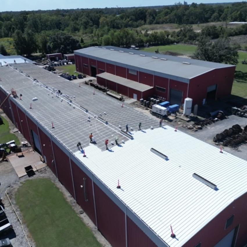 Aerial view of workers applying gaco silicone roof coating on a white metal roof atop a large red industrial building. The grassy surroundings and distant trees provide a picturesque backdrop, while equipment and materials are scattered around the site.