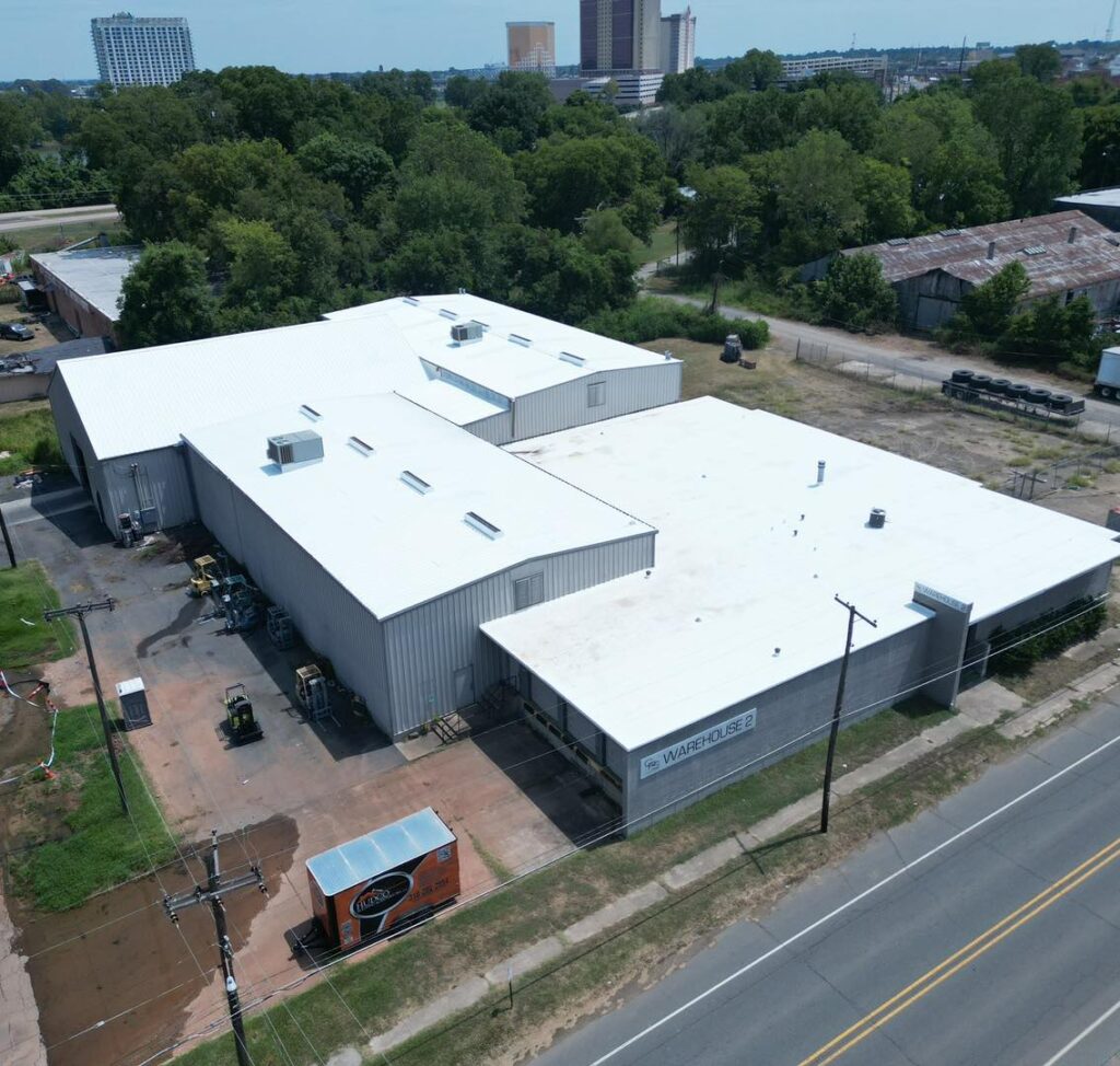 Aerial view of a large industrial building with a Gaco silicone-coated white roof, surrounded by greenery and a few other structures. A paved road runs in the foreground, and an orange container is visible near the building.