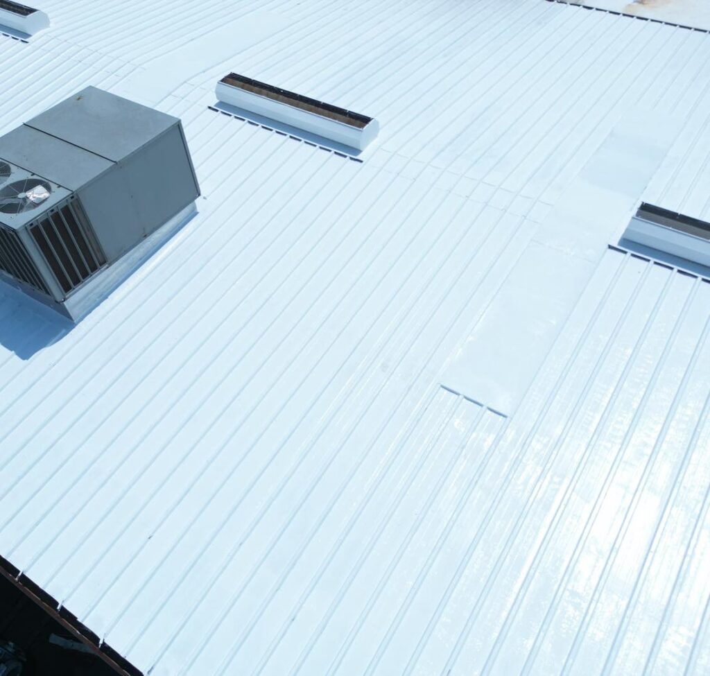 A metal rooftop with a white reflective Gaco silicone roof coating, featuring an air conditioning unit and skylights. This bright surface enhances energy efficiency and temperature control.