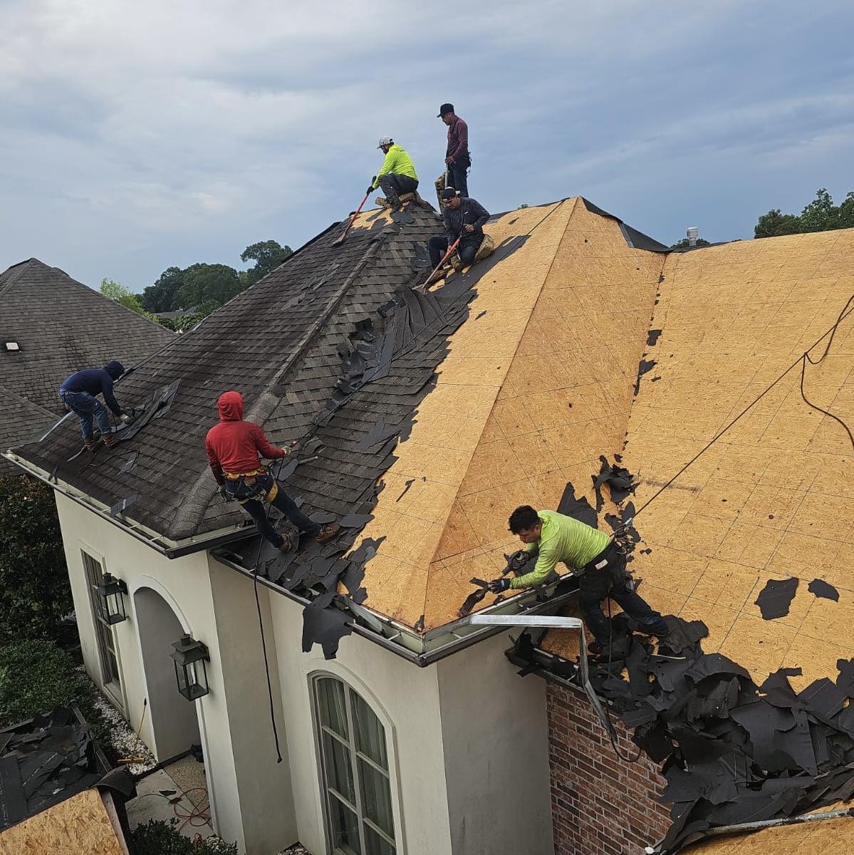 Workers on a steep roof are removing old shingles, contemplating roof repair vs replacement. Some are wearing safety harnesses. The sky is cloudy, and scattered tools are visible on the roof. The house is two-story with large windows and a mix of brick and stucco exterior surfaces.