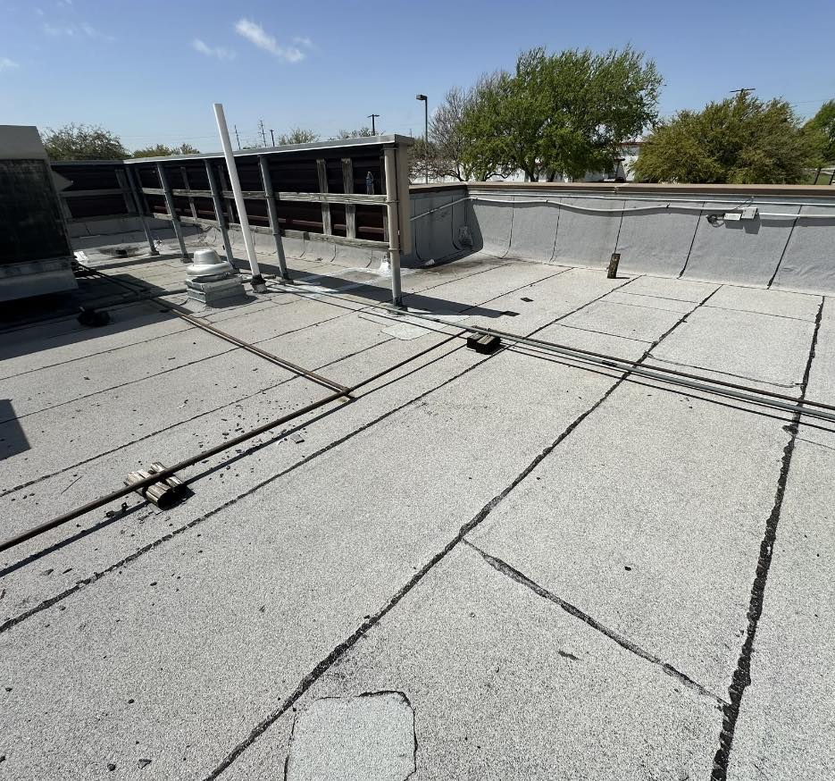 The flat rooftop, representative of Baton Rouge Commercial Roofing excellence, features a light gray surface. Several pipes and vents are visible alongside a low railing near the edge. In the background, trees stand tall against a clear blue sky, completing the serene scene.