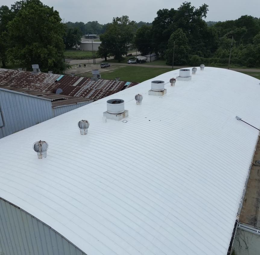 An aerial view showcases the expansive white industrial roof, a hallmark of Baton Rouge Commercial Roofing, with several ventilation units. Trees and other buildings stand in the background under a cloudy sky.