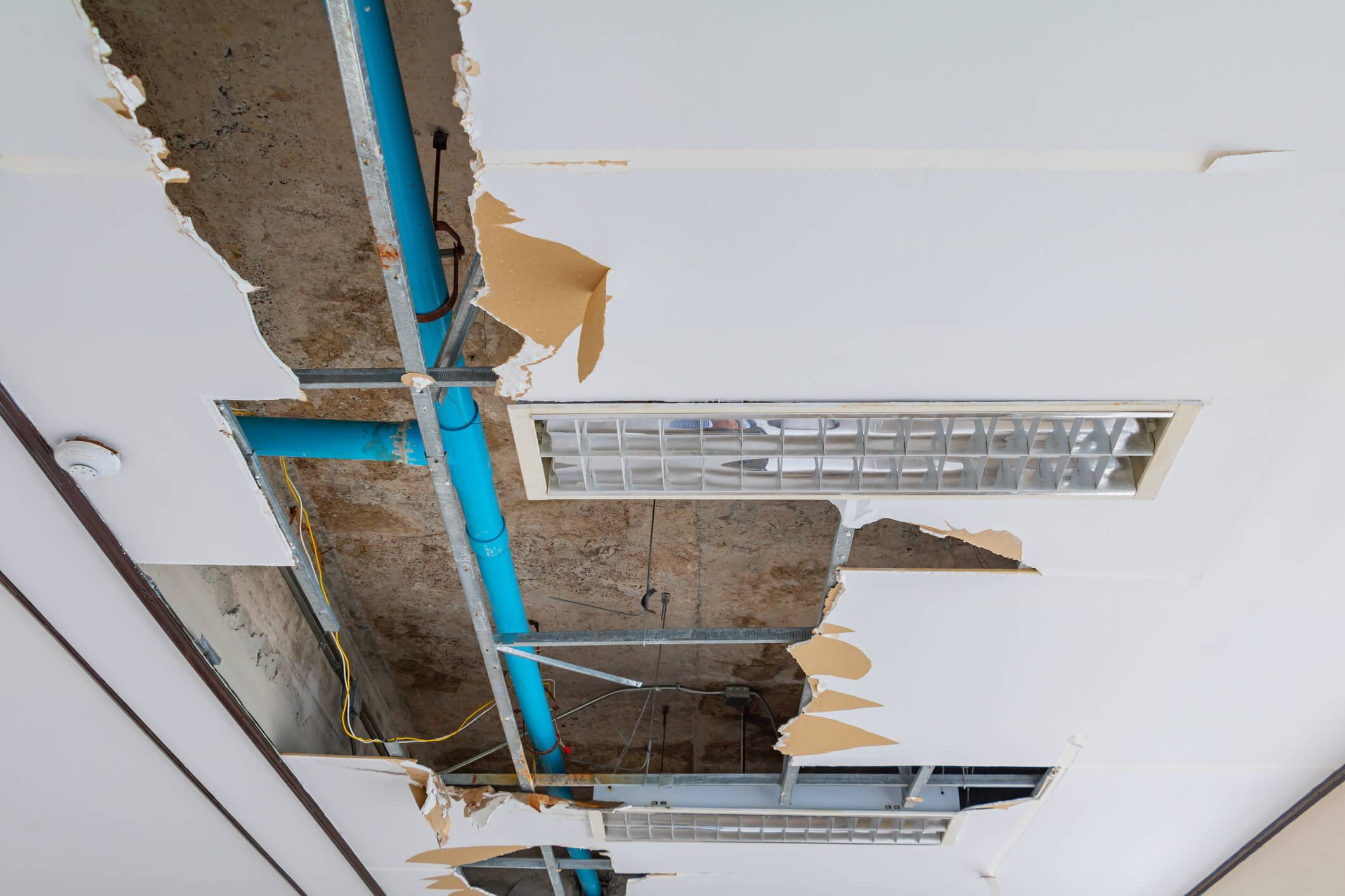 A commercial roof leak has led to a damaged ceiling with sections of plaster missing, revealing blue pipes, electrical wires, and metal ducts. A rectangular air vent is visible amidst the debris.