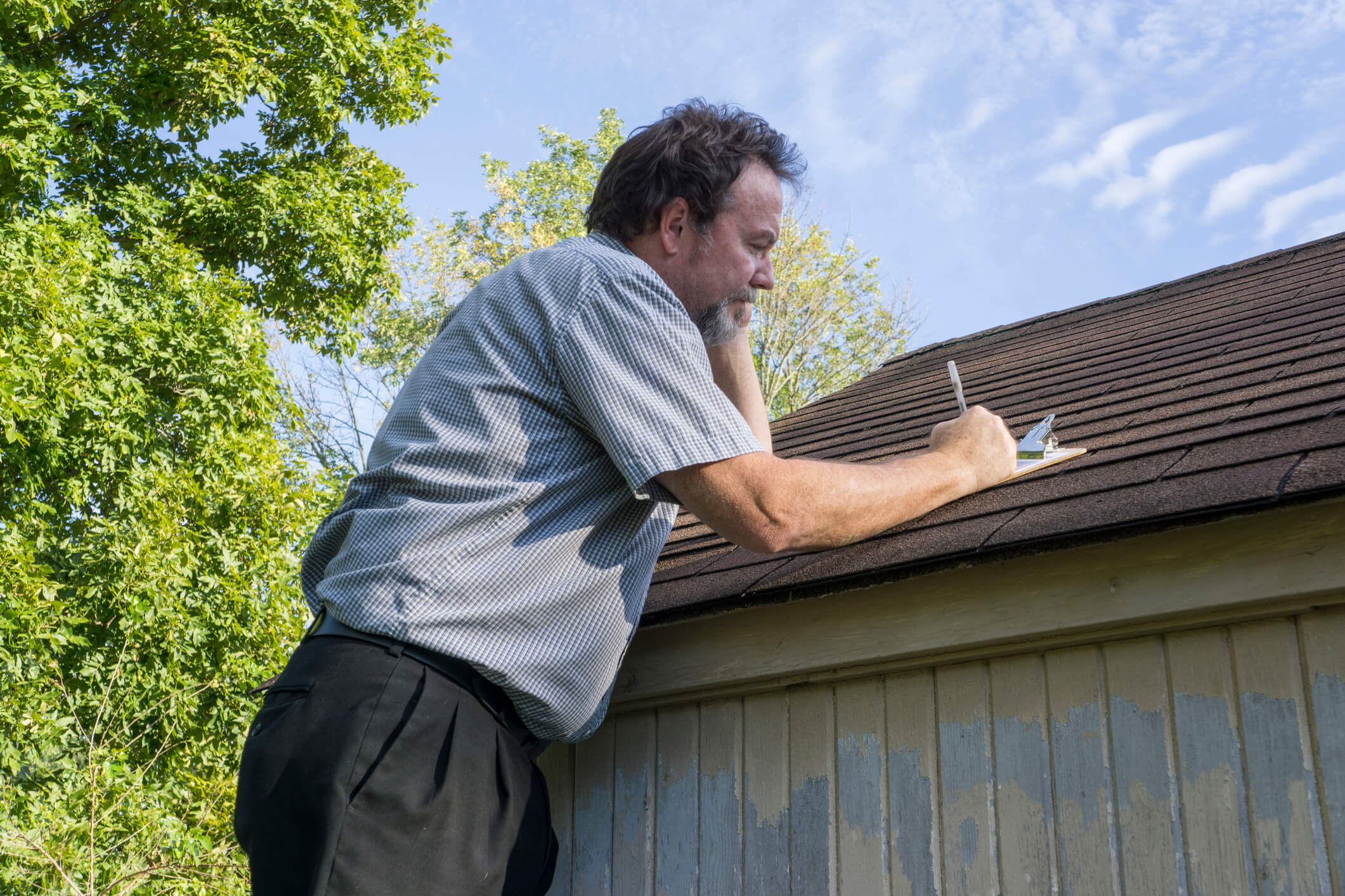 A man inspects the roof of a house, taking notes with a pen and notepad. He stands on a ladder in a short-sleeved shirt and pants, ready to address what to do if insurance denied a roof claim. Trees and a clear sky frame the scene.