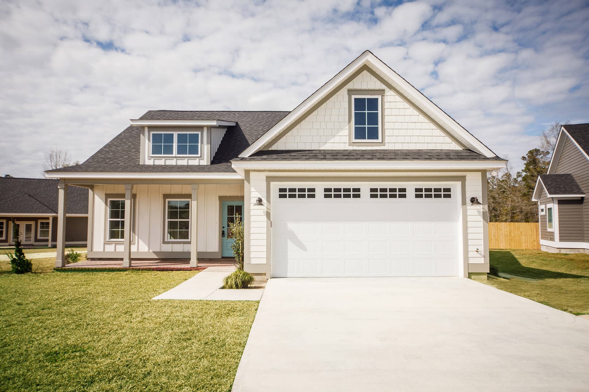 A modern suburban house with a light-colored exterior, a gabled roof, and a two-car garage invites contact. Featuring a cozy front porch and a neatly maintained lawn under a partly cloudy sky, this home beckons residents to enjoy its charm and comfort.