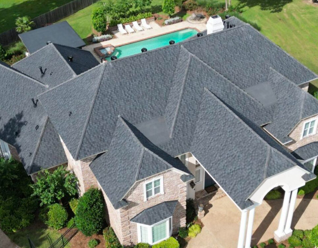 Aerial view of a large house with a grey, multi-gabled roof expertly crafted by a Louisiana roofing contractor. The property features a swimming pool with white lounge chairs in the backyard, surrounded by lush greenery. The house has brick and white accents, including columns at the entrance.