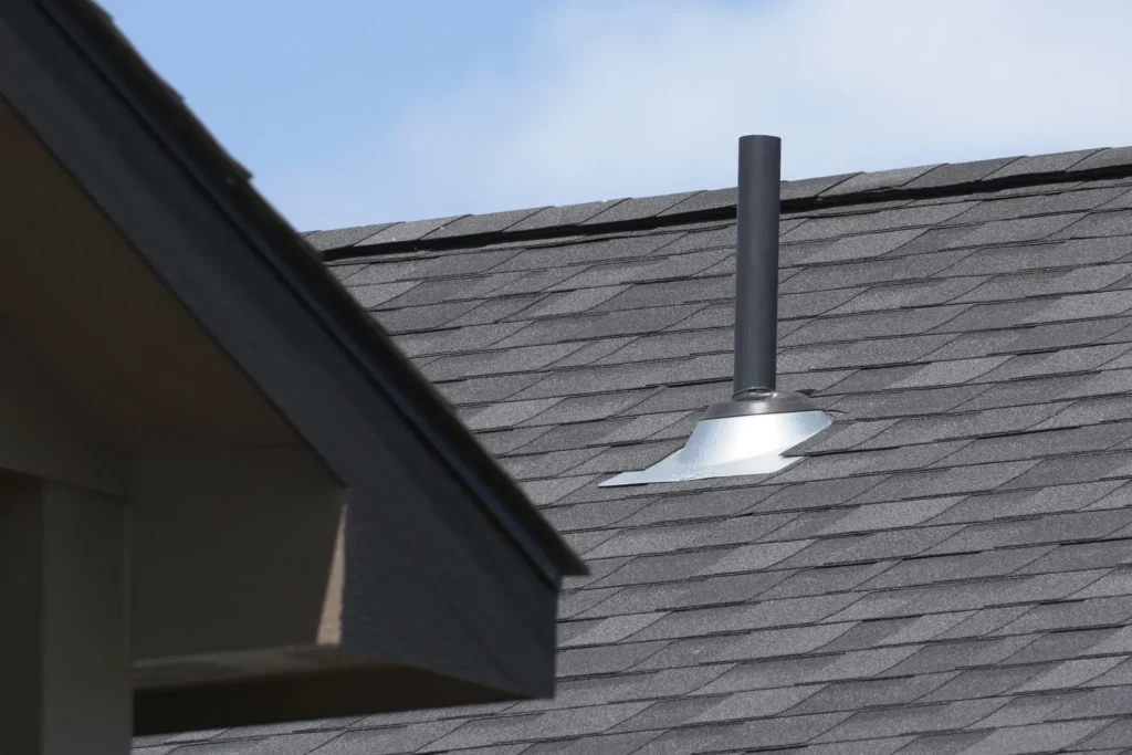 Under a clear blue sky, a gray shingled roof with a metal vent pipe stands resilient, as outlined in the 2025 Louisiana Roofing Guide. In the foreground, the edge of a neighboring roof adds depth to this quintessential view.