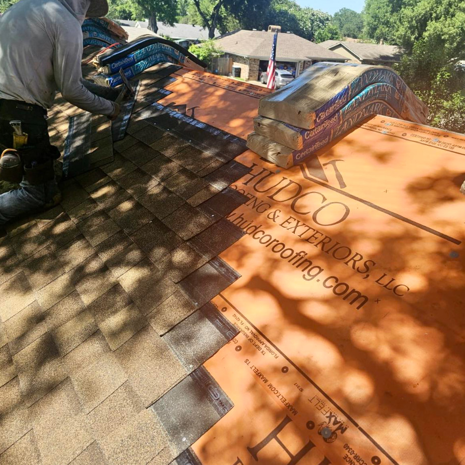 A person is installing shingles on a roof with visible roofing materials branded "Hudco Roofing & Exteriors, LLC." As the sun casts shadows from nearby trees onto the roof, skilled roofers bring their expertise to Baton Rouge, LA. In the background, houses and trees complete the scene.