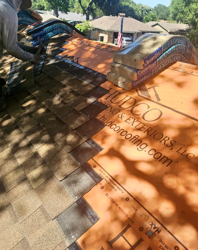 A person from a roofing company in Shreveport, LA, works diligently on a roof under construction, laying shingles atop the underlayment. The sunlight casts tree shadows on the surface while stacks of materials and glimpses of a residential neighborhood can be seen nearby.