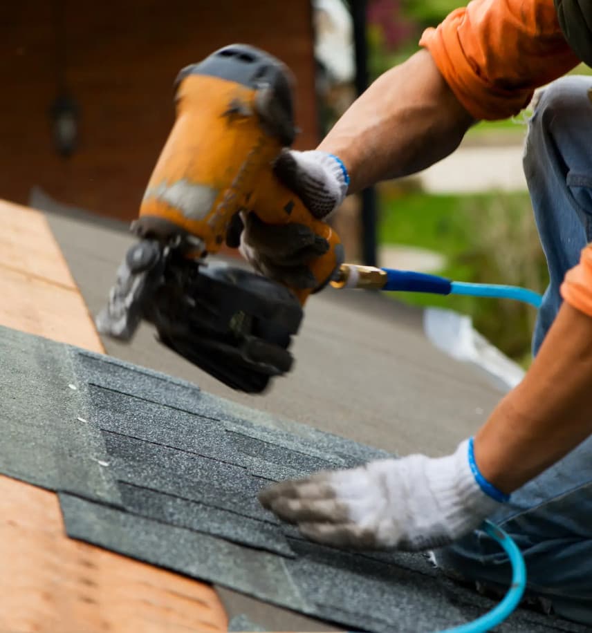 Close-up of a roofing contractor in Haughton, LA, using a nail gun to secure shingles. The individual is wearing an orange shirt and gloves. In the background, a blurred view reveals a brick wall and lush greenery.