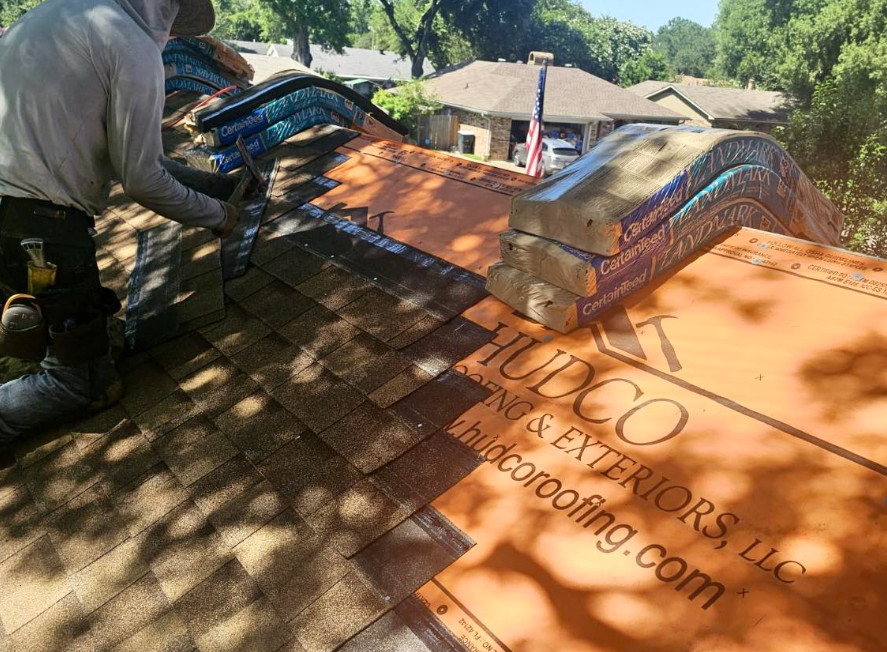 A roofing contractor in Lake Charles, LA, is installing shingles on a roof. The roof is partially complete, with orange underlayment visible. Bundles of shingles are stacked nearby, while trees and houses are visible in the background on a sunny day.