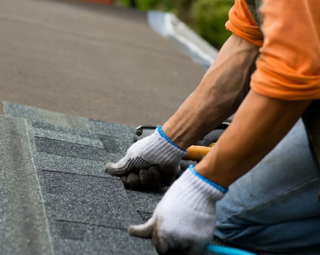 A roofing contractor in Thibodaux, wearing gloves and an orange shirt, is busy installing asphalt shingles on a sloped roof. Nearby, a tool lies ready for use amidst the dark gray shingles. In the background, lush greenery enhances the serene setting.