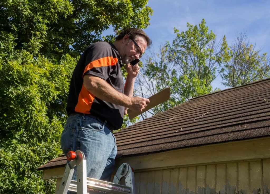 A man stands on a ladder beside a roof, talking on the phone and holding a clipboard. Dressed in a black and orange shirt with jeans, he's clearly overseeing roofing in Texarkana. Trees and a clear sky form the backdrop, signaling a sunny day perfect for outdoor work.