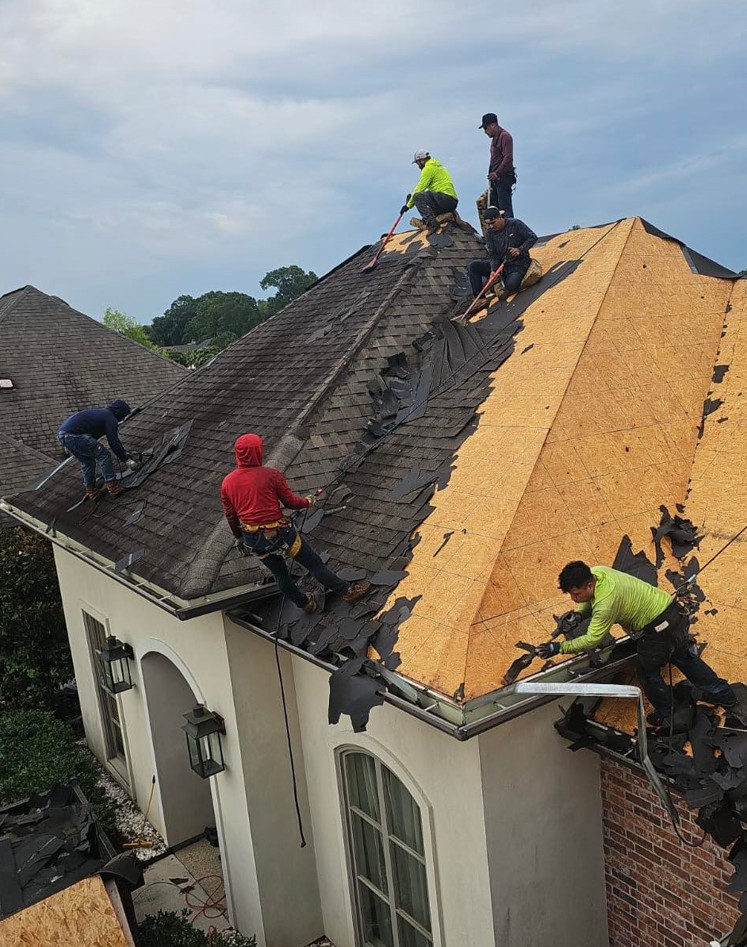 Five workers diligently remove old shingles from a steep residential roof in Baton Rouge, partially stripped down to the plywood. Clad in safety gear and bright clothing, they work under cloudy skies. The light-walled house with large windows and lanterns prepares for its upcoming roof insurance claims inspection.