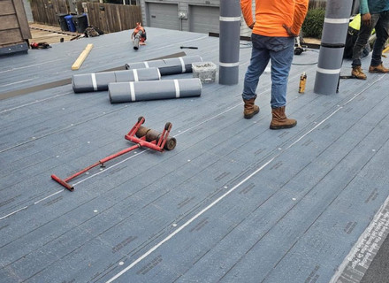 Workers on a rooftop installing rolled roofing materials. Some tools and equipment, including a red roller and roof rolls, are scattered on the surface. Two workers are standing, one in an orange jacket and the other in jeans.