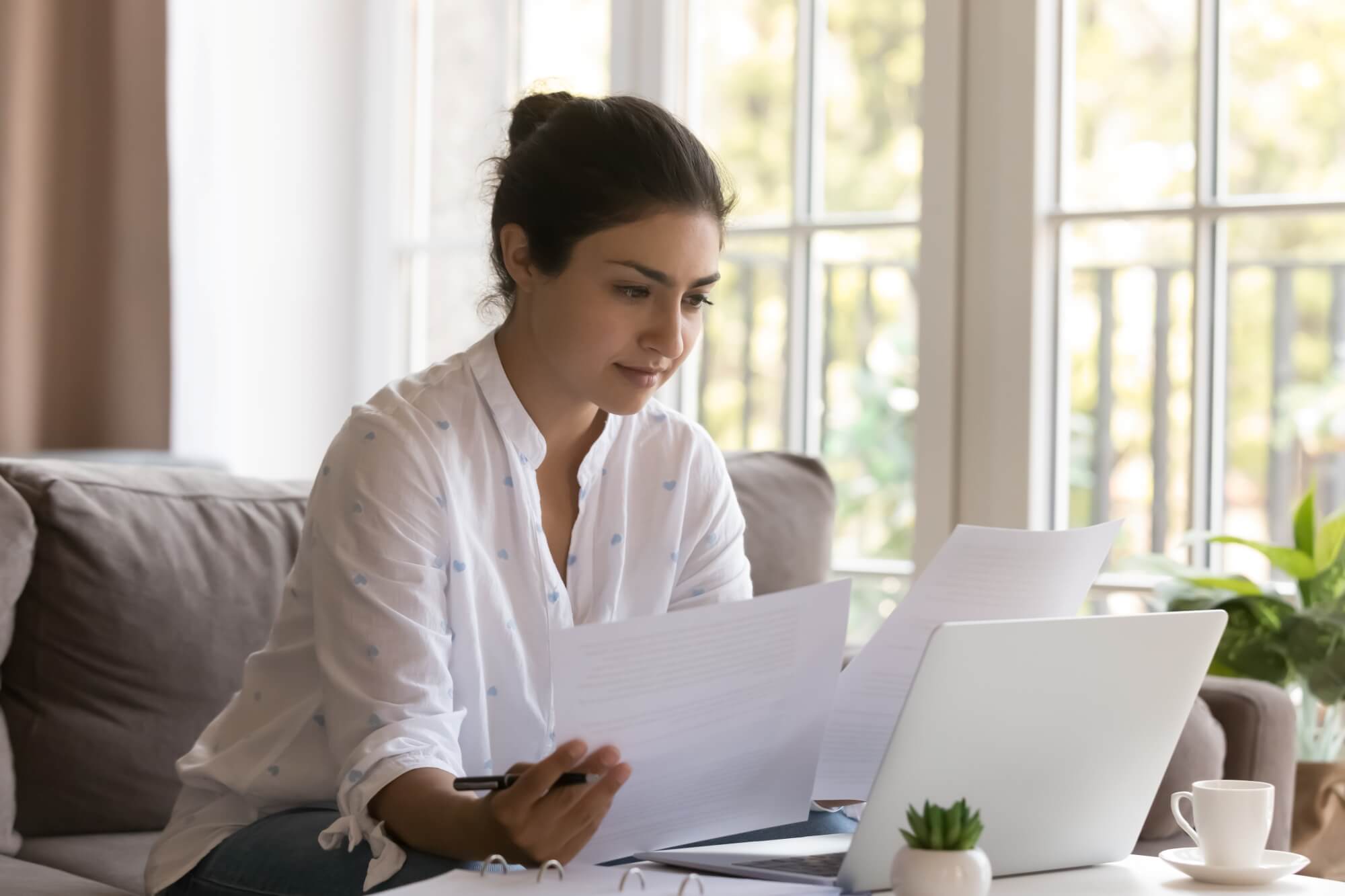 A woman sits on a sofa in front of a laptop, researching how to choose a roofing contractor without getting ripped off. She holds papers and a pen, focused, with a cup, small plant, and large windows letting in natural light behind her.