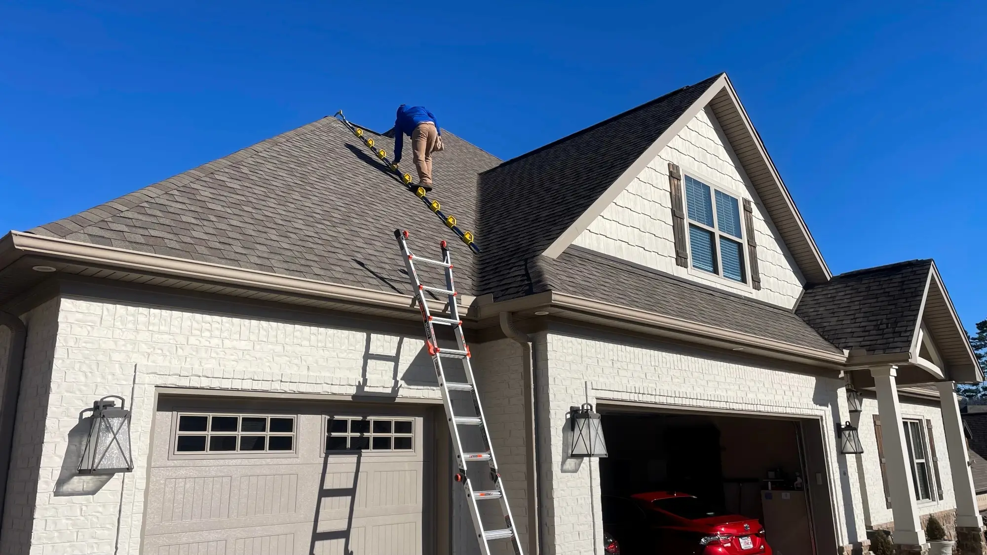 A person stands on a ladder placed on the steep roof of a beige two-story house, with another ladder leaning against the garage—a reminder of why learning how to choose a roofing contractor without being ripped off is important. The sky is clear and blue.