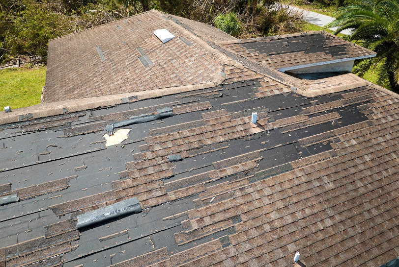 A house roof with missing and damaged asphalt shingles, exposing the black underlayment beneath. Several shingles are scattered or misaligned, indicating storm or wind damage. Trees and a road are visible in the background.